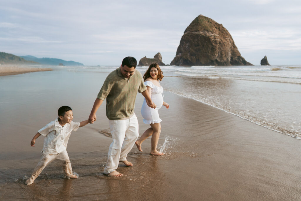 Family runs through the ocean water in Cannon Beach, Oregon while documenting their pregnancy with maternity photos