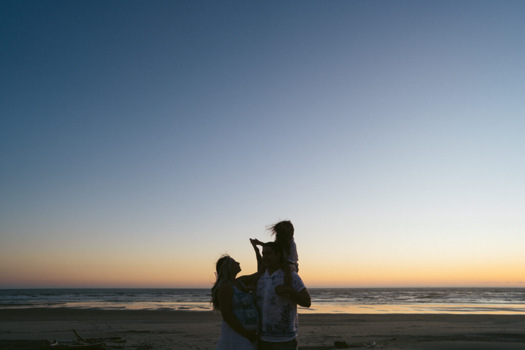 family stands on the beach on the Oregon Coast as the sun sets behind them and blue hour falls over the coastline. 